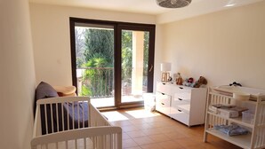 Ein heller Raum mit einem weißen Kinderbett, einer weißen Kommode und einem Balkon mit Blick auf Grünflächen.