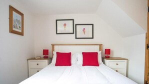 A bedroom with a white bed, two red pillows, a wooden headboard, two bedside tables with red lamps, and two framed pictures on the wall. - Abbey Place Net Loft (Penzance)