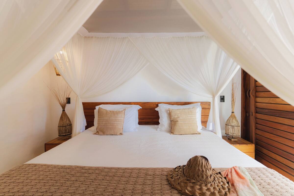 A bedroom with a large bed, white bedding, and a canopy. There are two bedside tables with lamps and a vase on one.