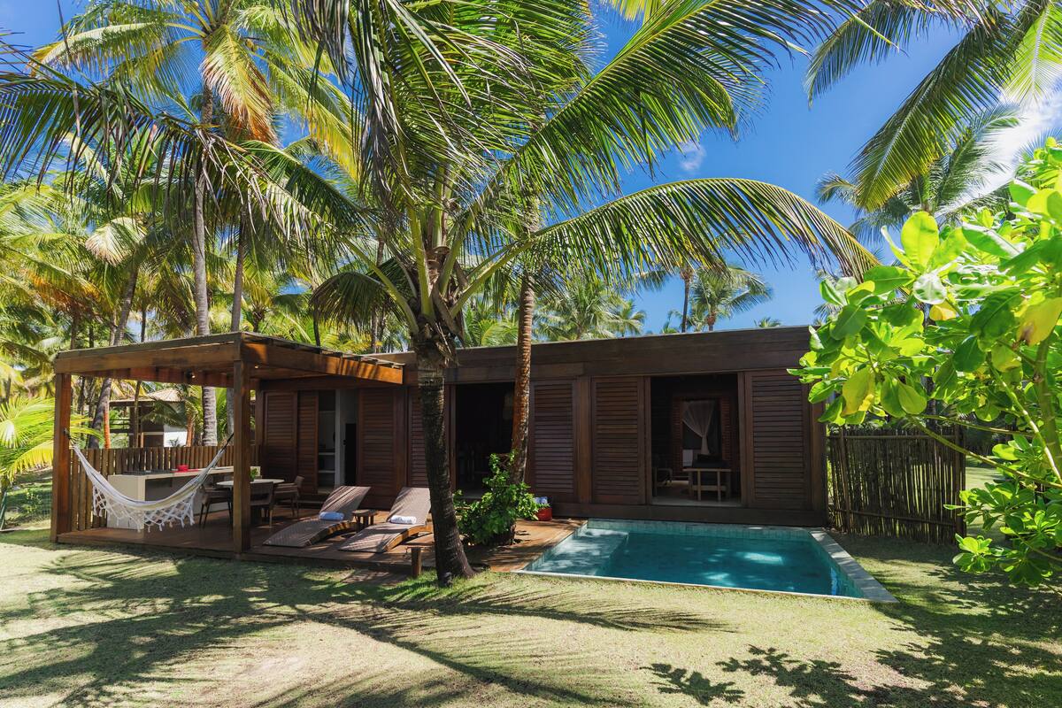 A wooden house with a pool, surrounded by palm trees and a hammock.