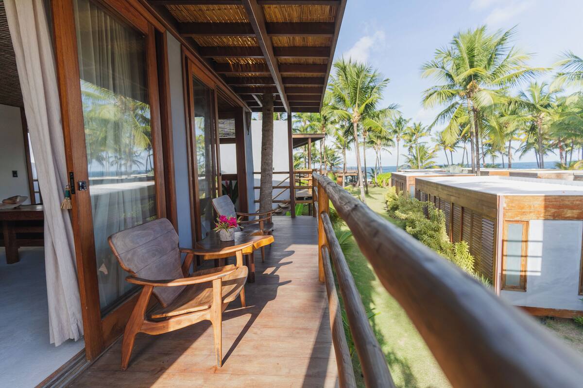 A wooden deck with chairs and a table, overlooking a beach with palm trees.