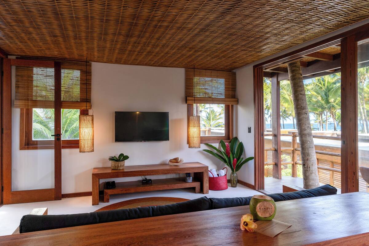 A modern living room with a flat-screen TV, wooden furniture, and large windows with views of palm trees.