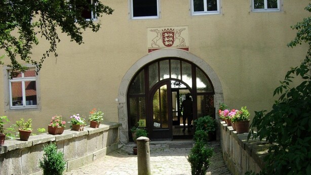 A cobblestone pathway leading to a building with a large arched entrance, potted plants, and a stone wall. - Hotel Schloss Sindlingen (Jettingen)