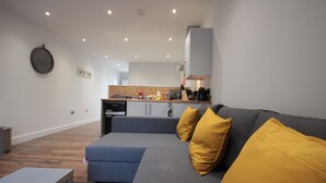 Uma sala de estar moderna com um sofá cinzento, almofadas amarelas e uma mesa de centro de madeira. Uma cozinha é visível ao fundo, com um painel de backsplash de tijolos e eletrodomésticos modernos.