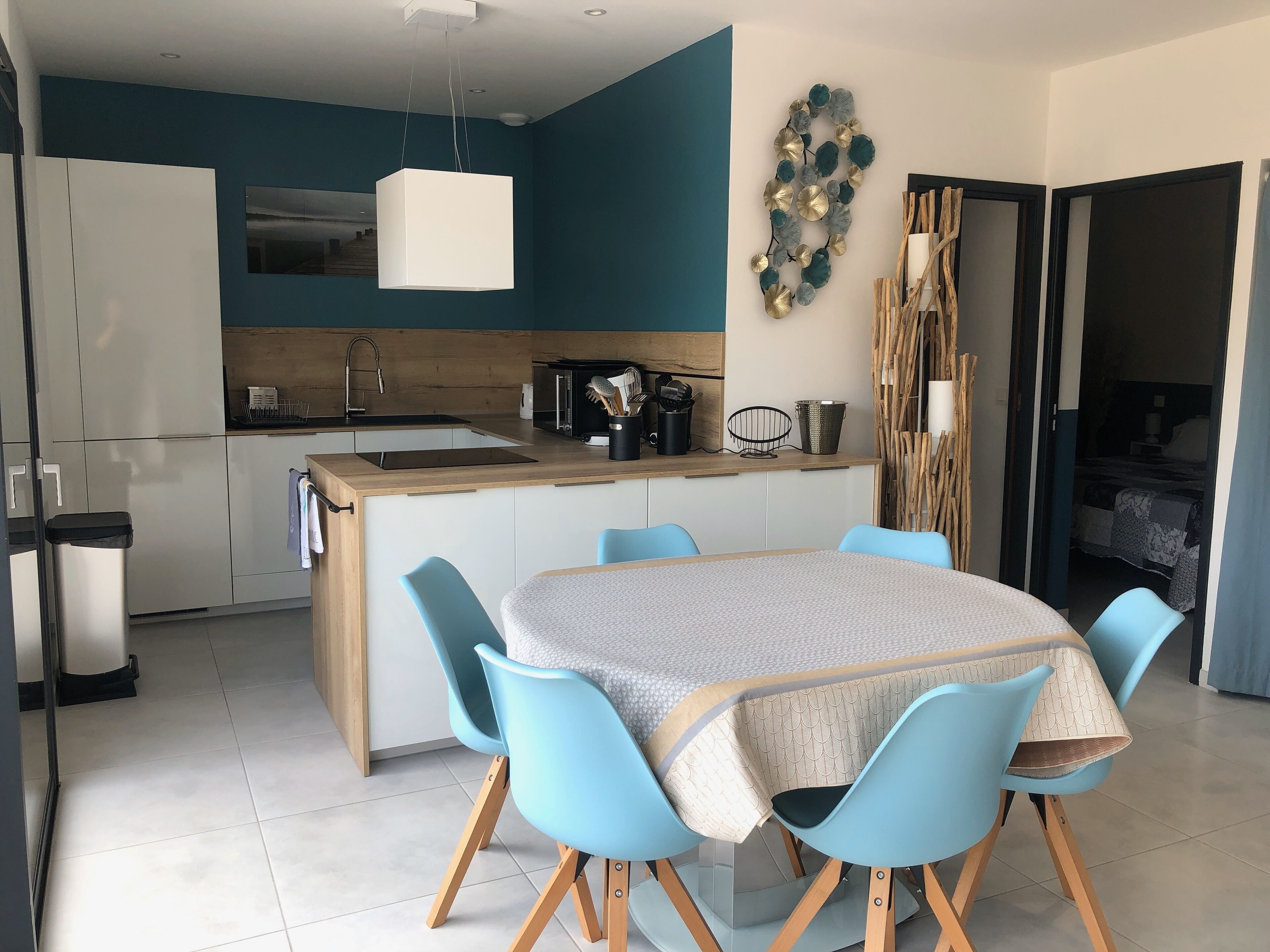 A modern kitchen with a central island, a dining area with blue chairs, and a living area with a bed and curtains.