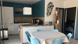 A modern kitchen with a central island, a dining area with blue chairs, and a living area with a bed and curtains. - Cala Rossa: MT **** charm + Quality / Price – Private heated pool-Air conditioning- WIFI (Lecci)