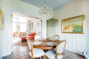 Uma sala de jantar com mesa de madeira, cadeiras acolchoadas brancas, um candelabro, um armário verde e uma pintura de paisagem.