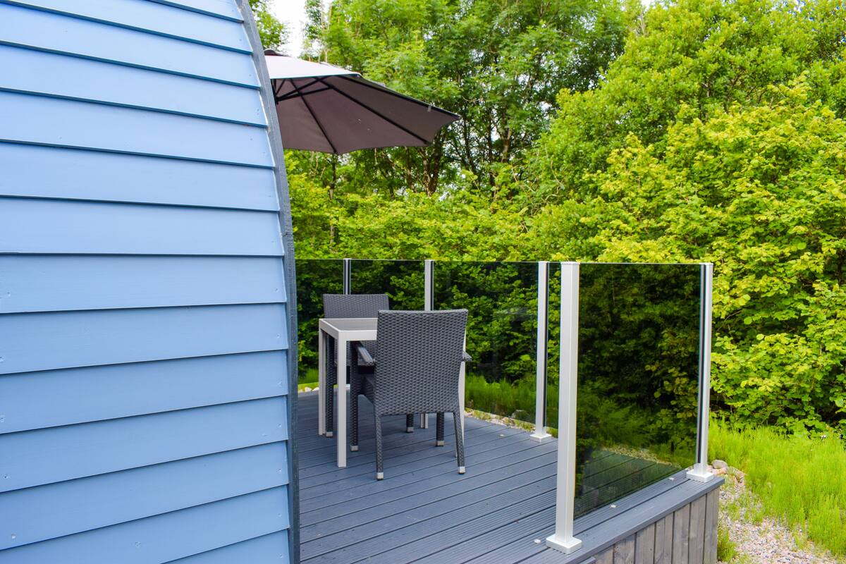 A deck with a table and chairs, surrounded by a glass railing, overlooking a lush green landscape.