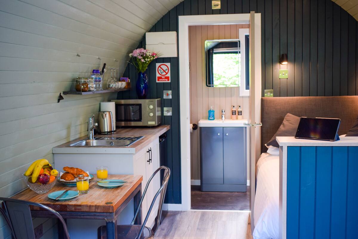 A compact kitchen with a microwave, toaster, and sink. There is a dining area with a table set for two, and a bed with a laptop on it.
