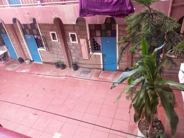 A building with a tiled ground floor and a balcony with a purple cloth. There are potted plants on the balcony and a large tree in the foreground.