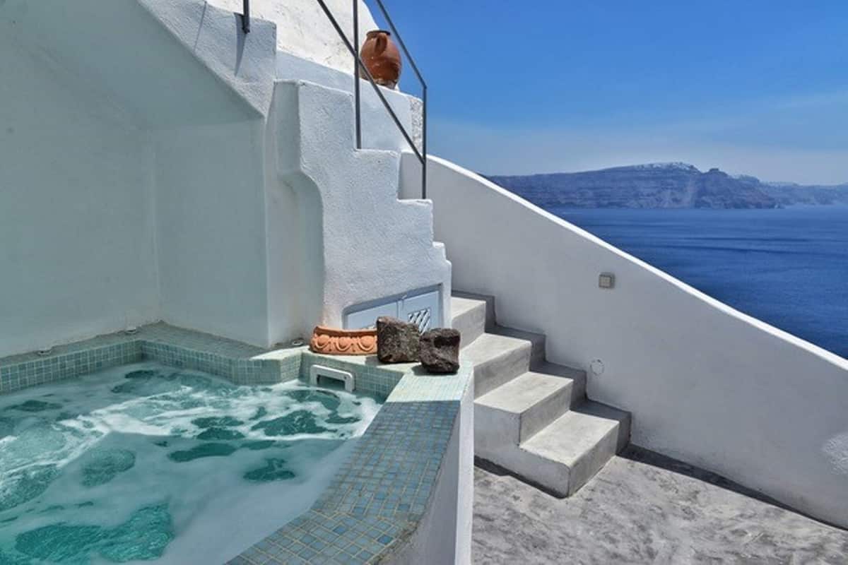 Una bañera de hidromasaje con vista al mar, paredes blancas y un camino de piedra que lleva a una puerta.