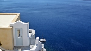 A coastal building with a white facade and a door, overlooking a deep blue sea with boats and distant landforms. - Utopia Villas - Amaya Selection Of Villas (Santorini)