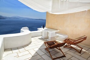 A sunlit outdoor seating area with a view of the sea, a white table with a bottle and glass, and two wooden lounge chairs. - Utopia Villas - Amaya Selection Of Villas (Santorini)