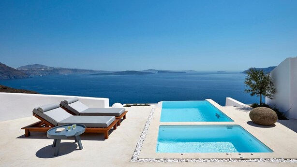 A pool area with lounge chairs and a small table, overlooking a clear blue sea and distant islands. - Utopia Villas - Amaya Selection Of Villas (Santorini)