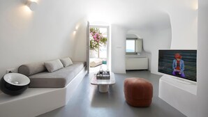 A modern living room with a sofa, a coffee table, and a television. There is a window with a view of a garden and a white wall. - Utopia Villas - Amaya Selection Of Villas (Santorini)