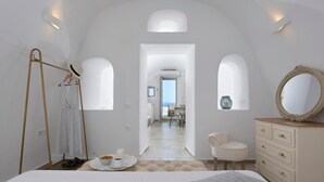A bedroom with a bed, a dresser, a chair, and a view of a balcony and dining area. - Utopia Villas - Amaya Selection Of Villas (Santorini)