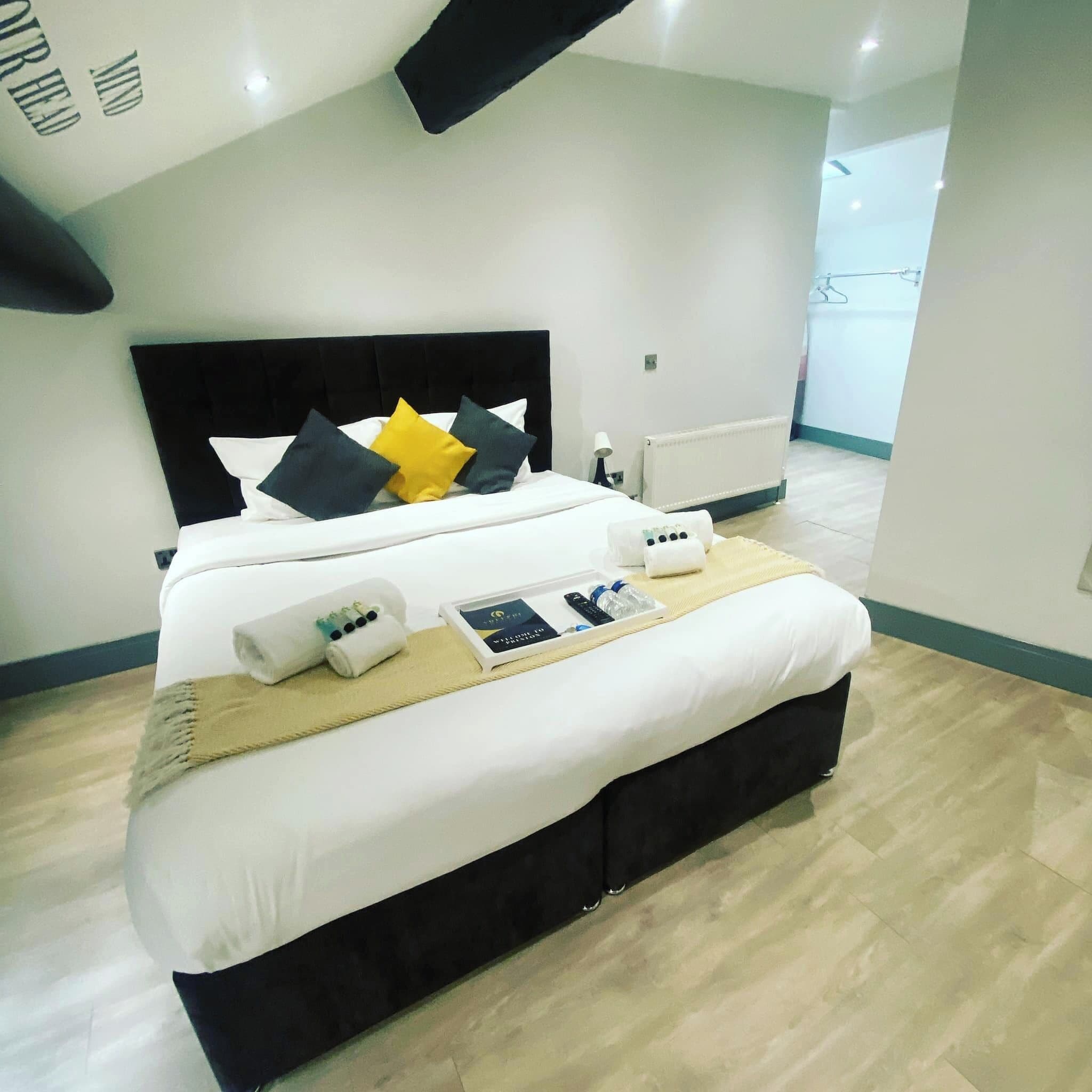 A neatly made bed with a black headboard, white bedding, and a few pillows in different colors. There is a tray with toiletries and a remote control on the bed.