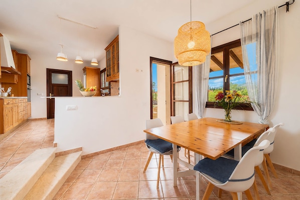 A dining area with a wooden table and chairs, a hanging light fixture, and a view through a window with curtains.