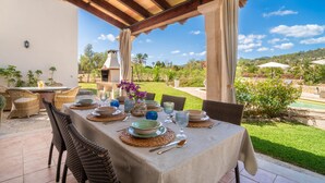 Un espace de restauration en plein air avec une table dressée pour quatre personnes, entourée de chaises en osier, sous une pergola en bois.