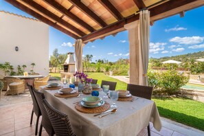 Un espace de restauration en plein air avec une table dressée pour quatre personnes, entourée de chaises en osier, sous une pergola en bois.