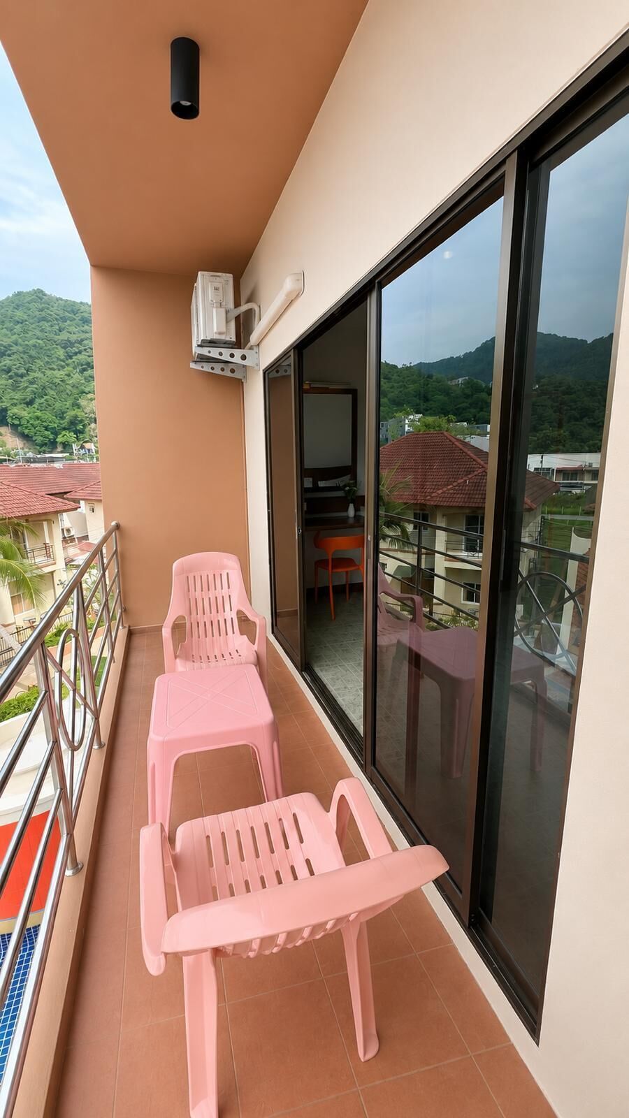 Un balcon avec des chaises en plastique roses et une table, une grande porte vitrée, et une vue sur une montagne et des immeubles.