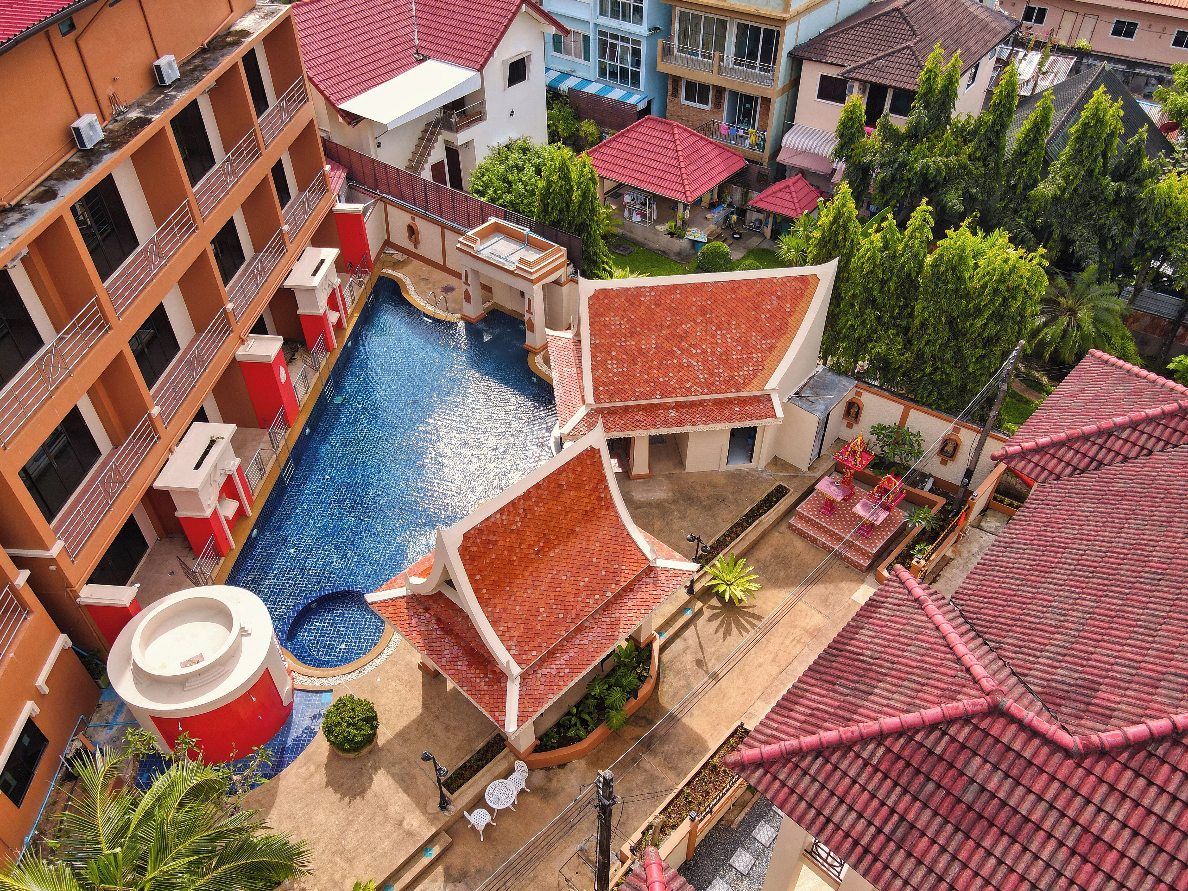An aerial view of a hotel with a central swimming pool, multiple balconies, and surrounding greenery.
