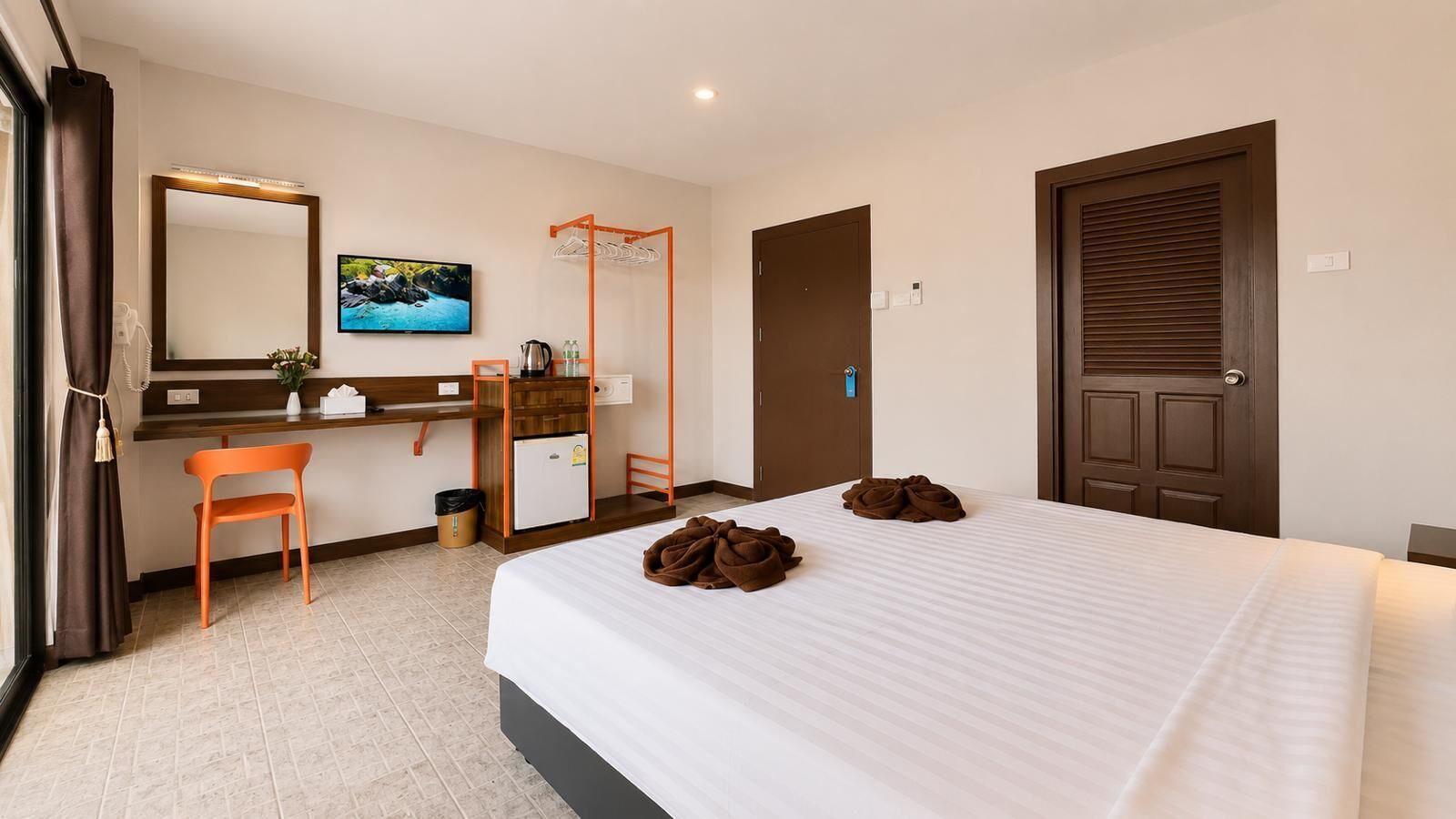 A hotel room with a bed, a desk, a chair, a small refrigerator, a mirror, and a door.
