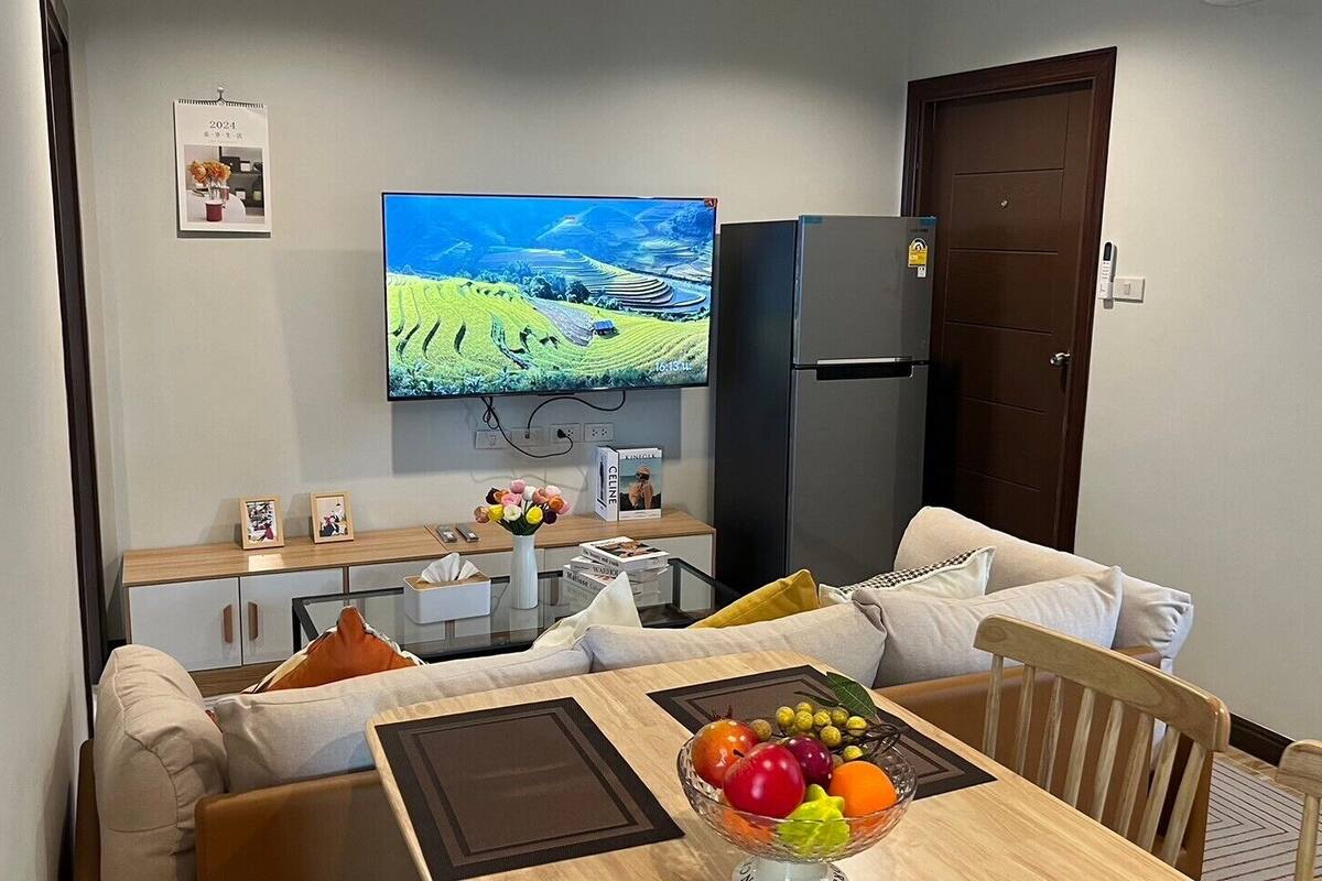 A modern living room with a flat-screen TV mounted on the wall, a dining area with a wooden table and a fruit bowl, and a kitchen area with a refrigerator and a wooden cabinet.