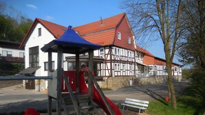 Ein Spielplatz mit Rutsche und Klettergerüst, im Hintergrund ein Haus mit rotem Dach und im Vordergrund eine Bank.