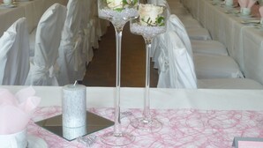 Ein Festsaal mit gedeckten Tischen für eine formelle Veranstaltung, mit weißen Tischdecken, rosa Servietten und Kerzenhaltern aus Glas mit Rosen.