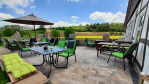 Eine Terrasse im Freien mit grünen Stühlen und Tischen, einem Grill und einem großen Sonnenschirm.