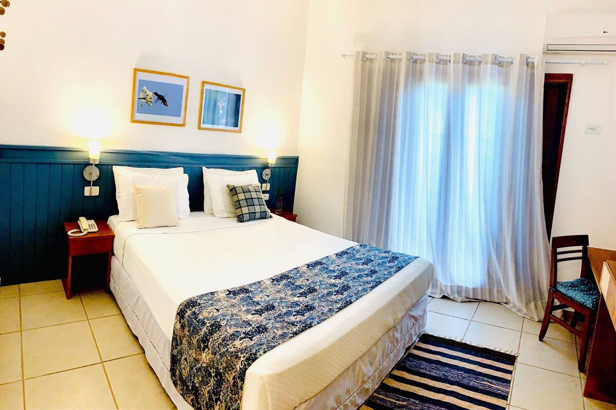 A neatly made bed with a patterned bedspread, a bedside lamp, and a telephone on a nightstand. There is a chair with a cushion, a window with sheer curtains, and a wall-mounted air conditioning unit.