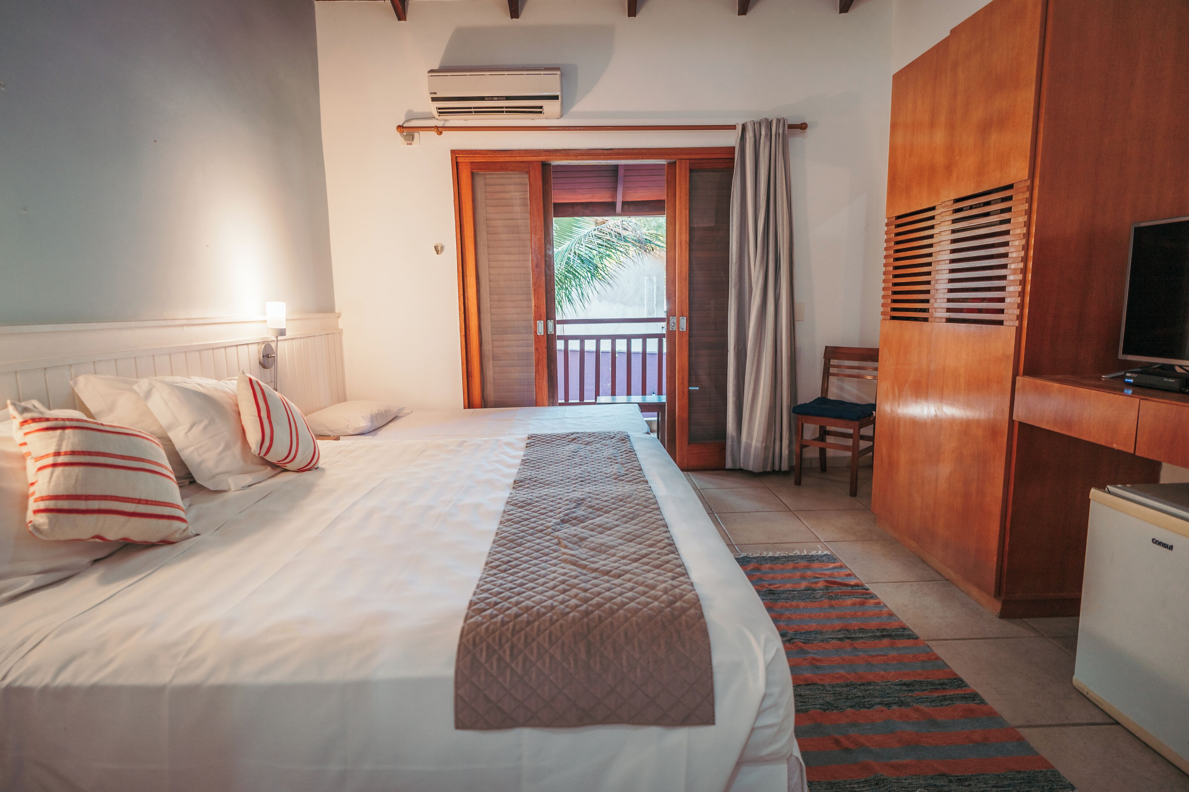 A hotel room with a bed, a television, a wardrobe, and a balcony with a view of palm trees.