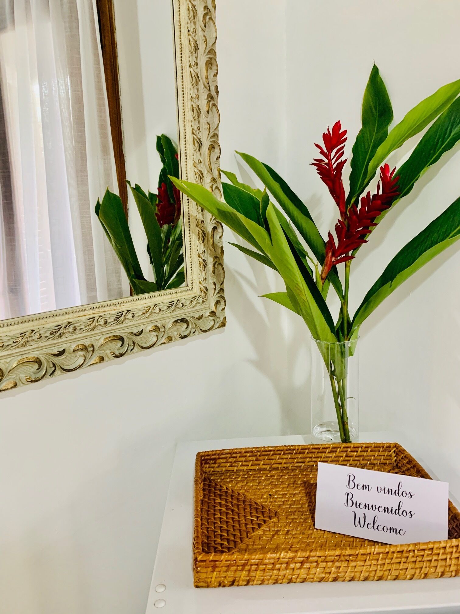 A decorative mirror with an ornate frame, a vase with red flowers and green leaves, and a welcome sign on a woven tray.