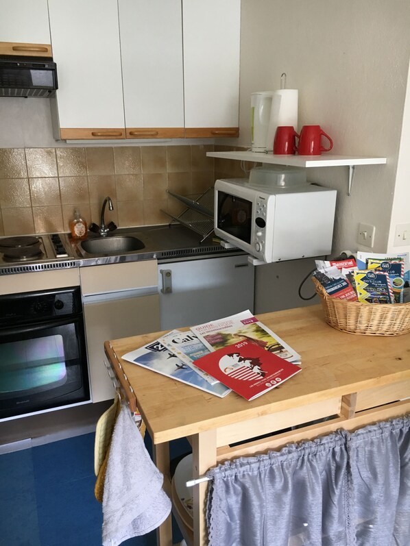 A compact kitchen with a microwave, stove, and oven. There is a sink, a small shelf with red cups, and a basket with snacks.
