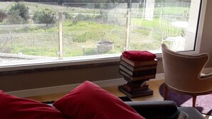 A bedroom with a large window offering a view of a lake and mountains. There is a bed with a quilt, a chair, and a stack of books on a side table. - escape escape from palermo (Piana degli Albanesi)
