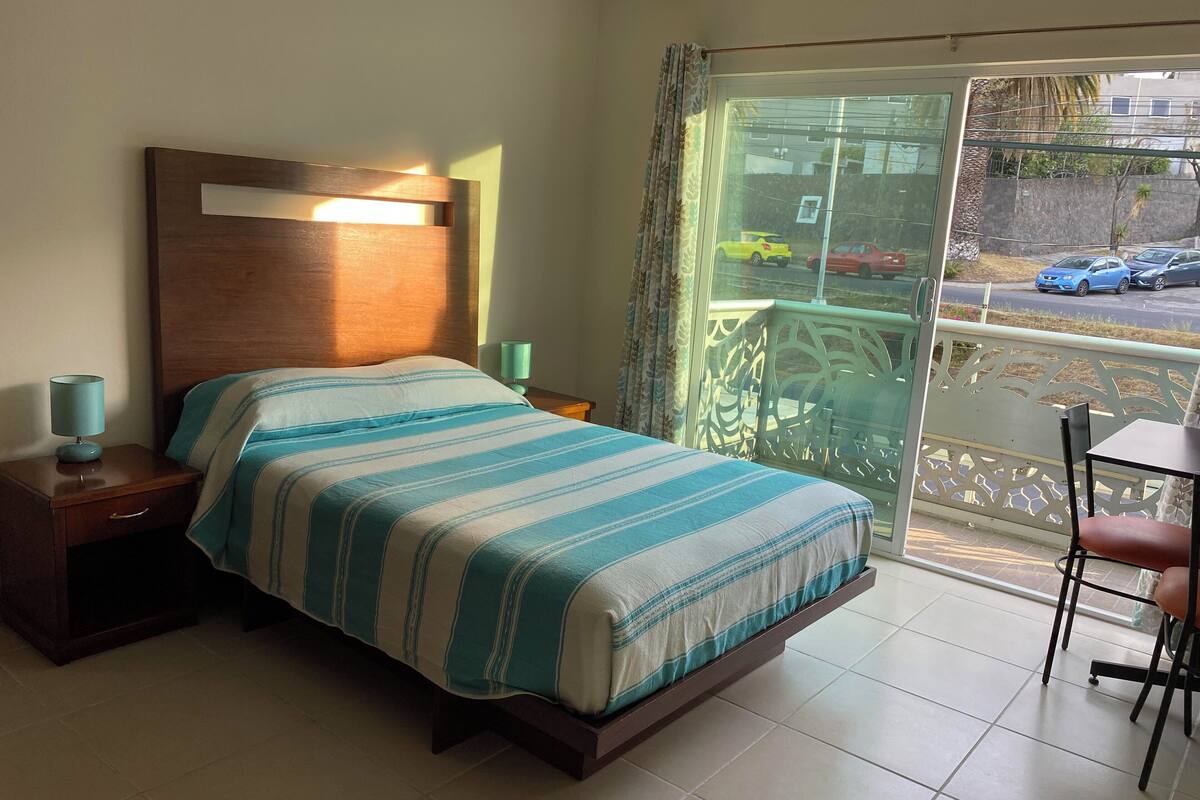 A hotel room with a bed, nightstands, a table, and a view of the street through a balcony.
