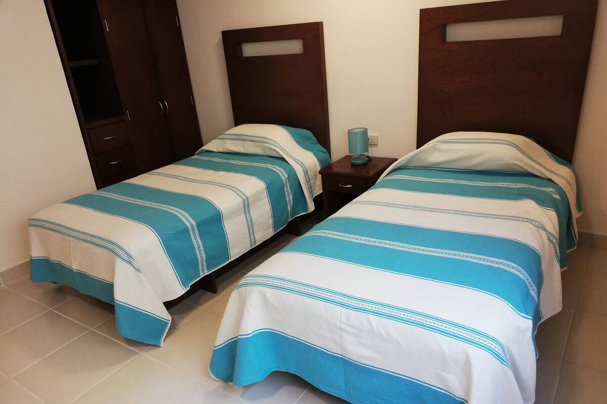 A room with two single beds, each with a blue and white striped bedspread, a small bedside table with a lamp, and a wooden wardrobe.