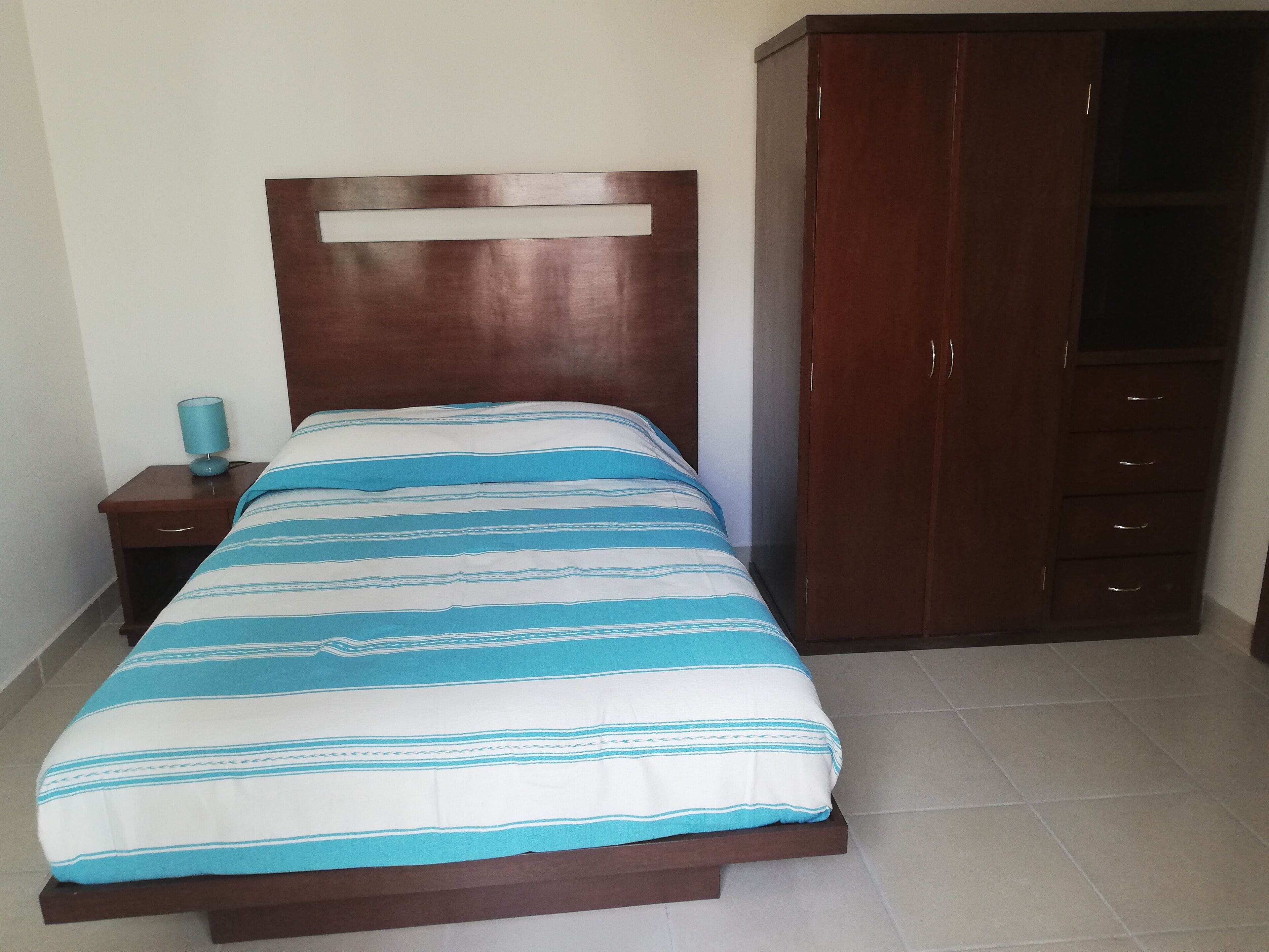 Un dormitorio con cama, mesita de noche, armario y una lámpara azul.