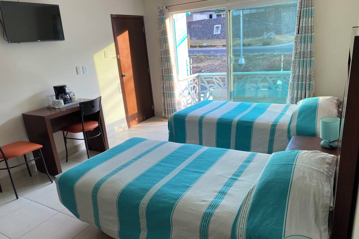 A hotel room with two beds, a desk with a chair, a TV mounted on the wall, and a balcony with a view of the outside.