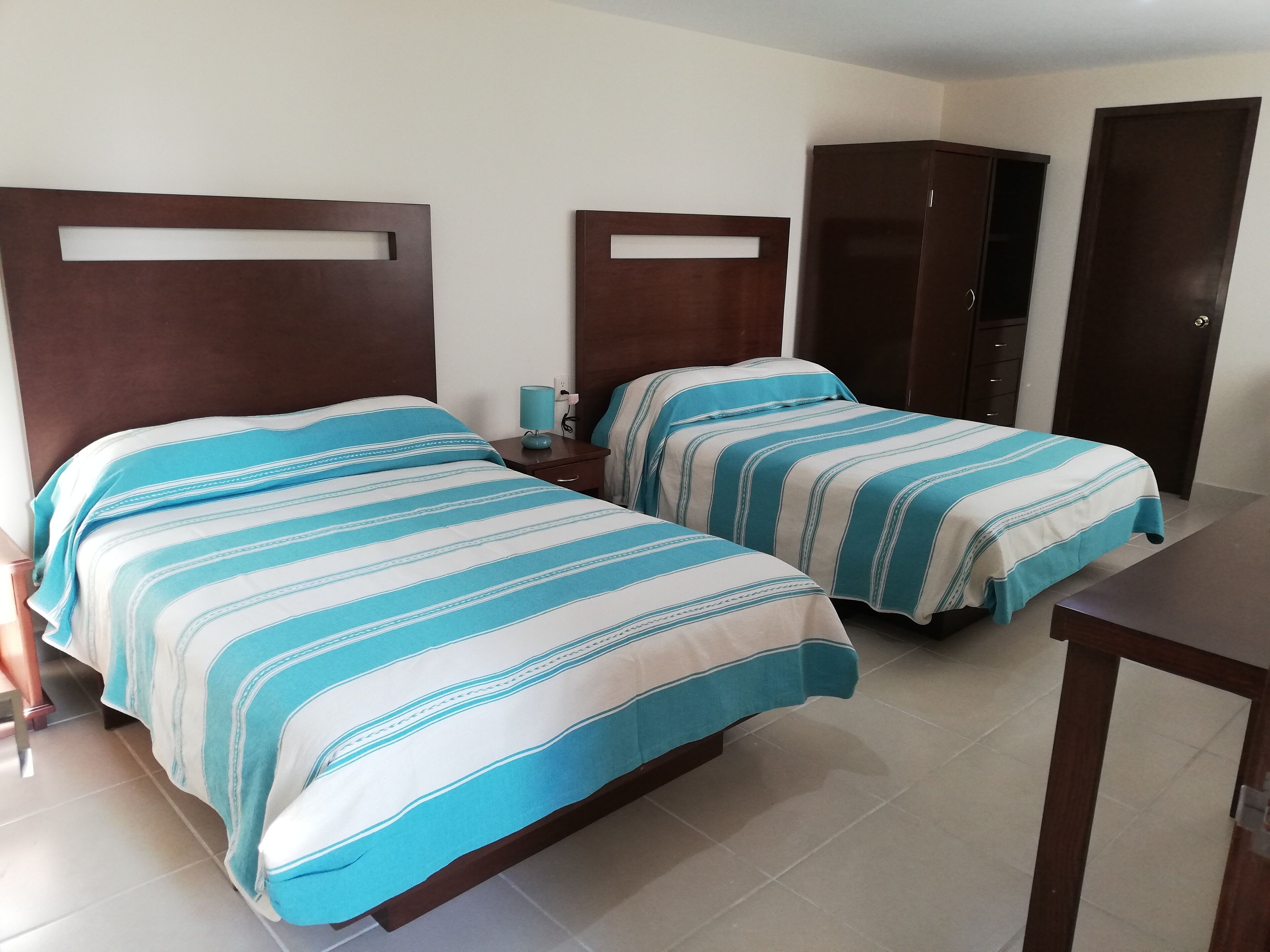 A hotel room with two beds, each with striped bedding, a bedside table with a lamp, and a wardrobe.
