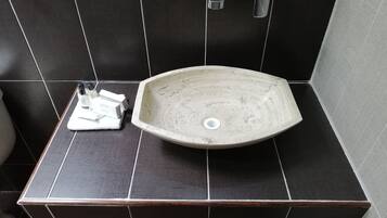 Een badkamer met een groot, ovaal keramieken wastafel, donkere betegelde muren, een houten aanrechtblad en een witte handdoek met toiletartikelen.