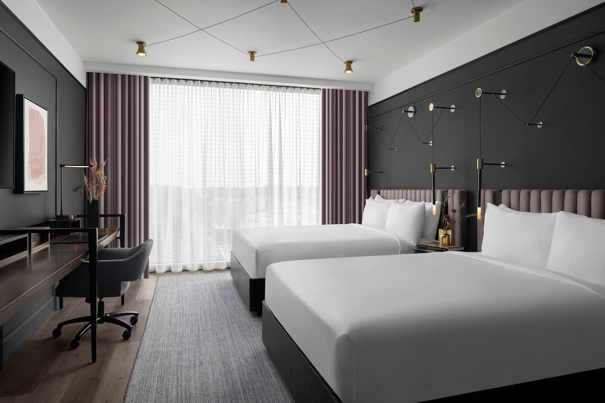 Ett modernt hotellrum med två sängar, ett skrivbord och ett stort fönster med gardiner.