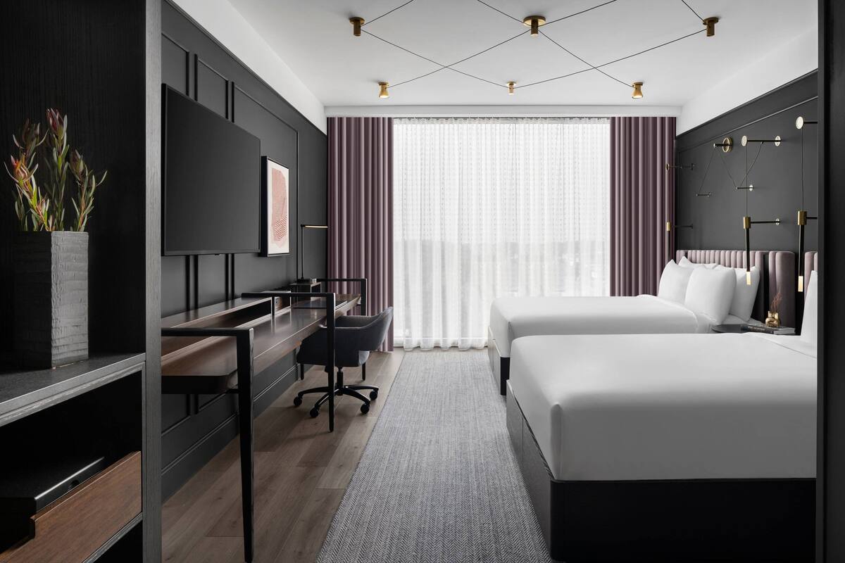 A modern hotel room with a large bed, a desk, a chair, a TV, and a large window with curtains.