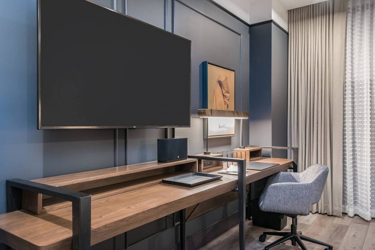 A modern hotel room with a wooden desk, a large flat-screen TV, a comfortable chair, and a large window with curtains.
