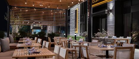 Un área de comedor al aire libre con mesas y sillas de madera, luces colgantes y un edificio con un letrero que dice "ELDR+RIME".