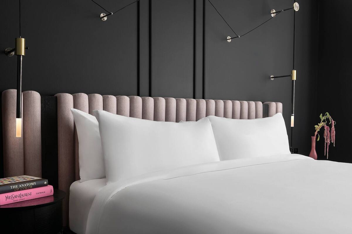 A neatly made bed with white linens, a dark headboard, and a bedside table with books and a vase.