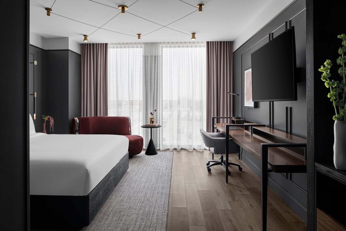 A modern hotel room with a large bed, a desk, a chair, a TV, and a plant.