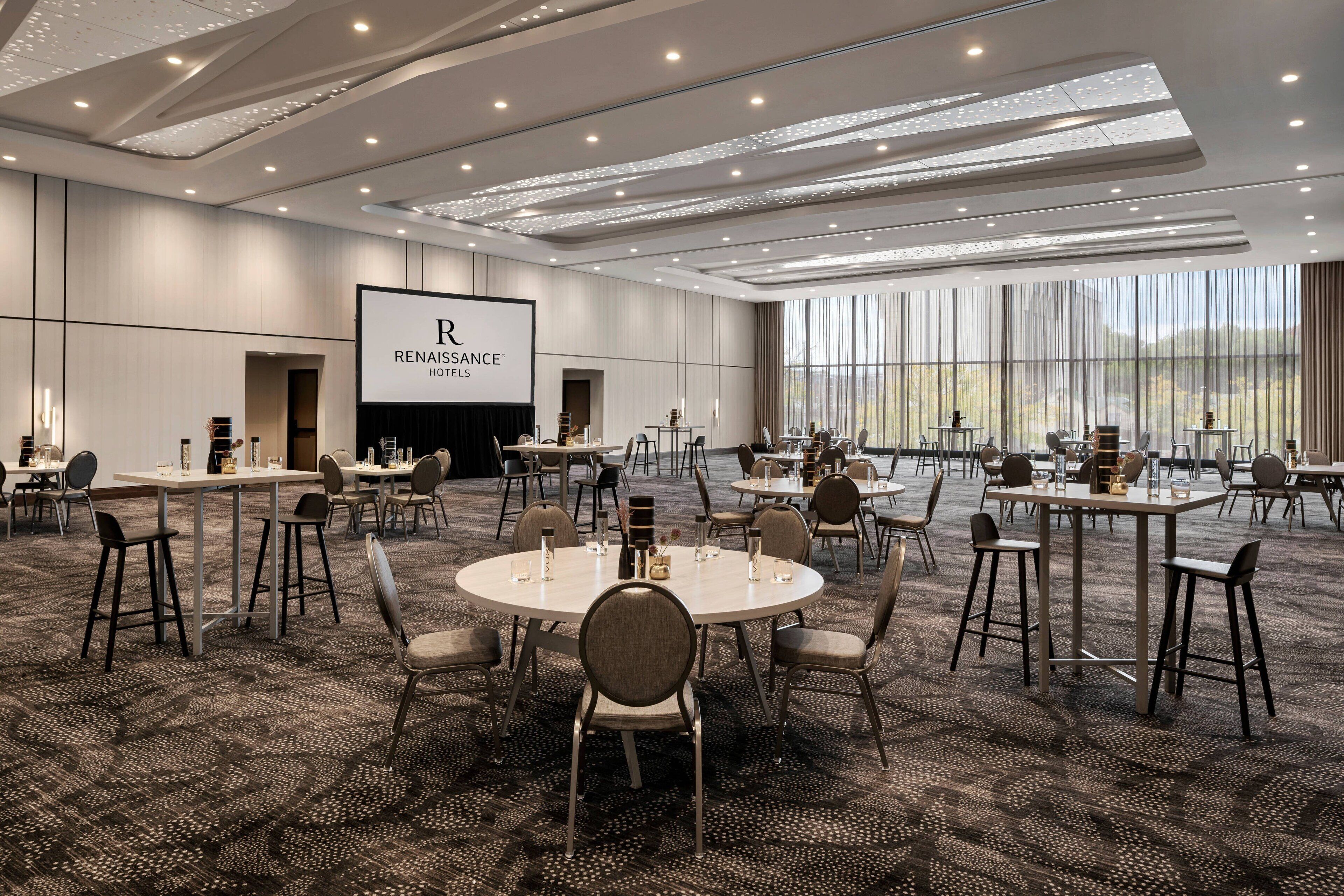 Un salón de conferencias con mesas y sillas redondas, una pantalla grande mostrando el logotipo del HOTEL RENAISSANCE, y grandes ventanales que ofrecen vistas al exterior.