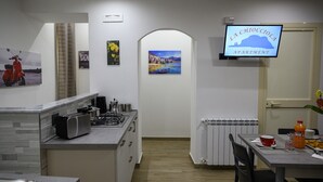 A compact kitchen with a stove, microwave, and cabinets, leading to a dining area with a table and chairs. A TV is mounted on the wall displaying 'LA CHIOCOLA APARTMENT'.
